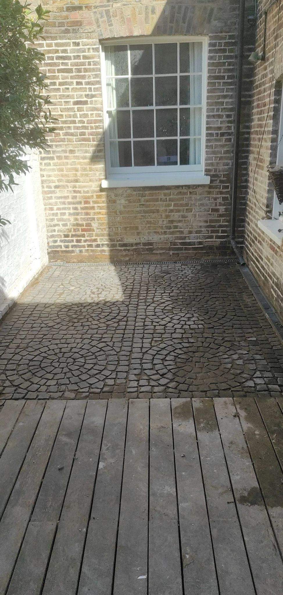 Decorative block paving patio featuring circular pattern in mixed tones with wooden decking and brick wall